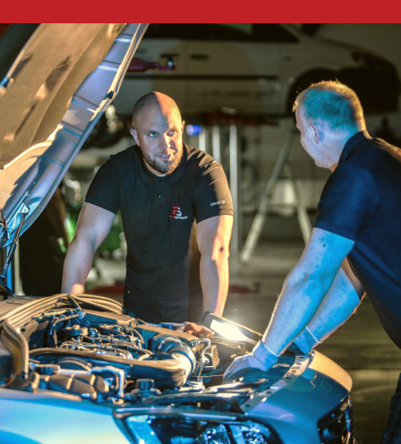 Two mechanics inspecting the engine of a car with the hood open inside a workshop.
